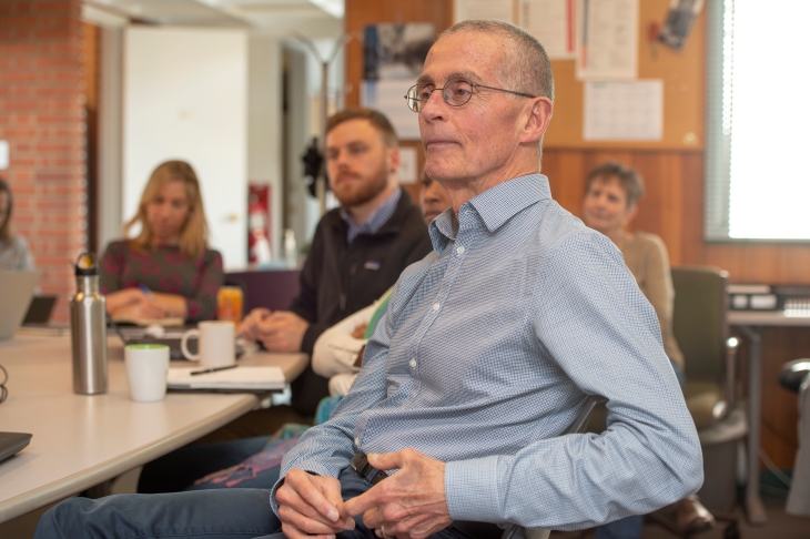 Man in glasses wearing blue shirt, listening attentively in meeting room, colleagues blurred