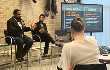 Two speakers on stools at a conference panel, audience member with bun and presentation screen