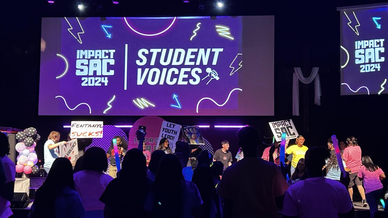 Purple-toned impact of Student Voices event with people in the foreground, signs, and event title slide in background.