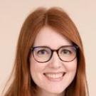 A smiling woman with long red hair and glasses, wearing a green sweater, against a neutral background.